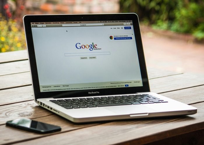 A MacBook Pro displaying Google Search on a wooden table outdoors, next to a smartphone.